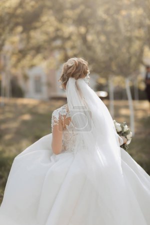 Portrait de mariage. Une mariée blonde en robe blanche avec un train marche, sourit et tient un bouquet et sa robe de mariée. Séance photo dans la nature. Rayons de soleil sur la photo
