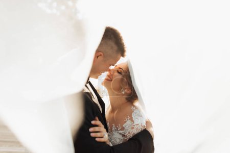 Wedding portrait. The groom in a black suit and the blonde bride are hugging, wrapped in the bride's veil. Long dress in the air. Photo session in nature. Beautiful hair and makeup