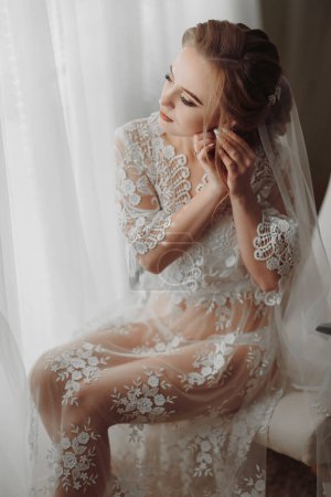 Téléchargez les photos : La mariée en robe transparente s'assoit sur le bord d'une chaise rose douce dans une pièce lumineuse, porte une élégante boucle d'oreille en or - en image libre de droit