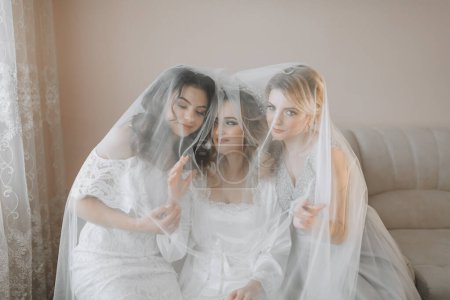 Photo for The bridesmaids look at the smiling bride. All on the same couch under a veil. The bride and her fun friends celebrate the bachelorette party in different dresses. Bride and friends in the room - Royalty Free Image