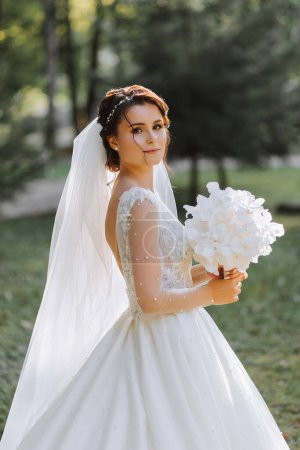 A cute bride in a lace dress is enjoying the holiday, standing in the park with a bouquet of white orchids. Portrait of the bride.