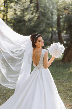 A cute bride in a lace dress is enjoying the holiday, standing in the park with a bouquet of white orchids. Portrait of the bride. Long white dress with open shoulders