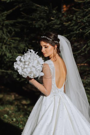 A cute bride in a lace dress is enjoying the holiday, standing in the park with a bouquet of white orchids. Portrait of the bride. Long white dress with open shoulders