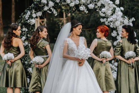 a beautiful bride of holds a wedding bouquet in her hands and looks at her bridesmaids who are holding bouquets with flowers behind their backs