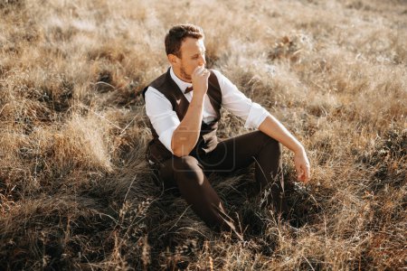 portrait of a stylish groom on a background of dry autumn grass. the concept of a rural wedding in the mountains, happy bohemian newlyweds. man relaxing sitting on the grass