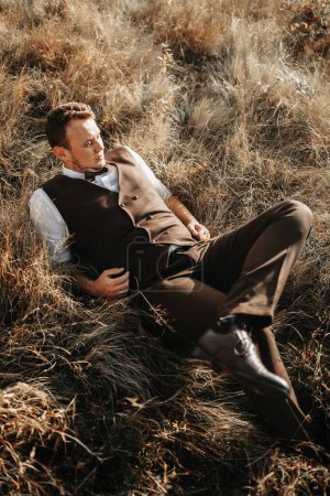Téléchargez les photos : Portrait d'un marié élégant sur un fond d'herbe sèche d'automne. le concept d'un mariage rural dans les montagnes, heureux jeunes mariés bohème. homme relaxant couché sur l'herbe - en image libre de droit