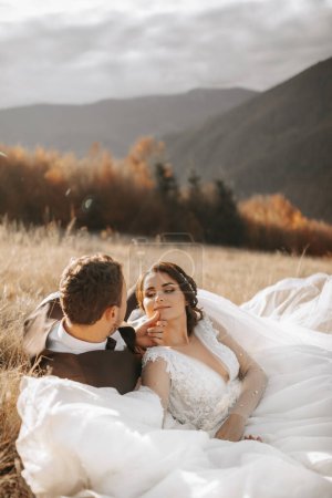 portrait of a stylish groom with a bride on a background of autumn dry grass. the concept of a rural wedding in the mountains, happy bohemian newlyweds. the bride and groom are lying on the grass