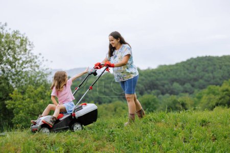 Foto de Una mujer en botas con su hijo en forma de un juego corta la hierba con una cortadora de césped en el jardín contra el fondo de las montañas y la niebla, concepto de herramientas de jardín - Imagen libre de derechos