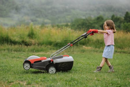 Foto de Un niño en botas en forma de un juego corta hierba con una cortadora de césped en el patio contra el fondo de las montañas y la niebla, el concepto de herramientas de jardín - Imagen libre de derechos