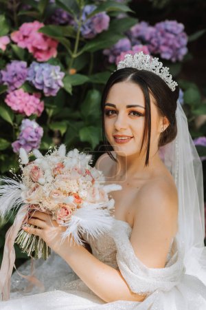 Photo for Young beautiful bride in wedding dress with open shoulders and crown on her head sitting near hydrangea flowers, fashion photo taken under harsh sunlight - Royalty Free Image