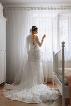 A beautiful young bride in a white wedding dress in a room near a window. Final preparations for the wedding. The bride is waiting for the groom. Morning, bride.
