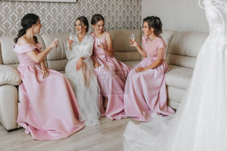 Téléchargez les photos : La mariée, avec des copines dans des robes roses identiques, le matin, assis sur le canapé, célébrer et se réjouir avec des verres de champagne dans leurs mains - en image libre de droit