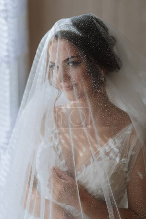 Die Braut trägt ein elegantes Kleid, das mit einem Schleier bedeckt ist, posiert und hält einen Boutonniere. Hochzeitsfoto, Morgen der Braut