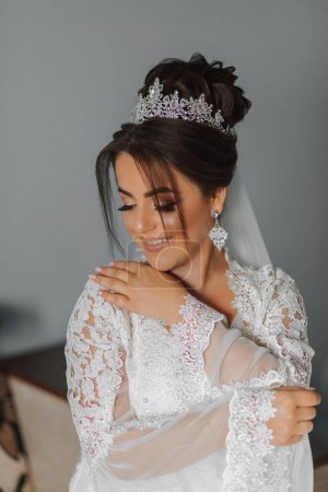 Téléchargez les photos : La timide mariée se couvre des manches de sa robe de dentelle. Portrait d'une mariée brune avec une tiare sur la tête et maquillage professionnel - en image libre de droit