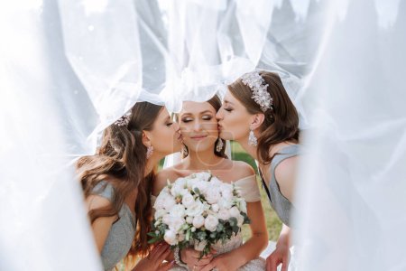 Téléchargez les photos : Des copines embrassent la mariée sur les joues, enveloppées dans un voile. La mariée tient un bouquet. Filles dans des robes identiques - en image libre de droit