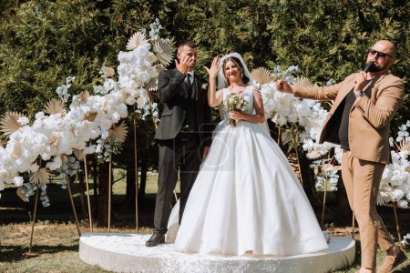 Téléchargez les photos : Cérémonie de mariage en nature. Les mariés près de la voûte florale. Maître de cérémonie dans des verres noirs lors d'un mariage lors d'une performance sur le fond de la mariée et le marié. - en image libre de droit