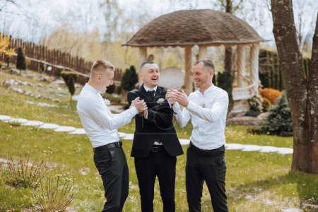 Three men in suits are standing in a grassy area, one of them is a wedding officiant