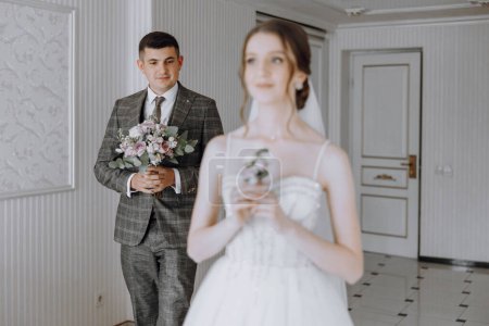 Un hombre y una mujer están en una habitación con una puerta blanca. El hombre sostiene un ramo de flores y la mujer sostiene un pequeño ramo. La escena es romántica e íntima
