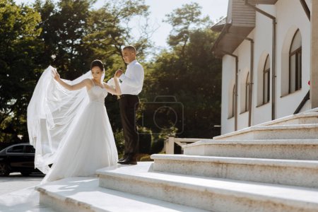 A bride and groom are standing on a set of white steps, with the bride holding a white veil