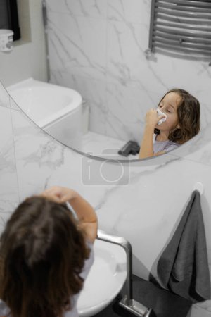 A young girl blows snot from her nose in front of a mirror