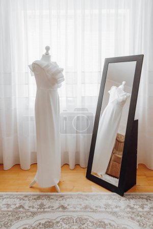 A white dress is standing in front of a mirror. The dress is long and elegant, and the mirror is black. The room is well-lit, and the dress is the main focus of the image