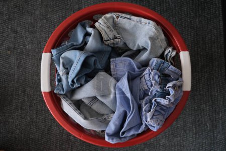 Photo for Denim pants, Clean unfolded denim trousers in a red basket on a yellow carpet. Disordered denim clothes concept idea photo. Top view of a red basket full of denim clothes. - Royalty Free Image