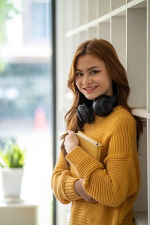 Photo for Young Beautiful smiling happy student woman wearing headphone holding a book, Portrait of student girl carry book balancing books on head and hand - Royalty Free Image