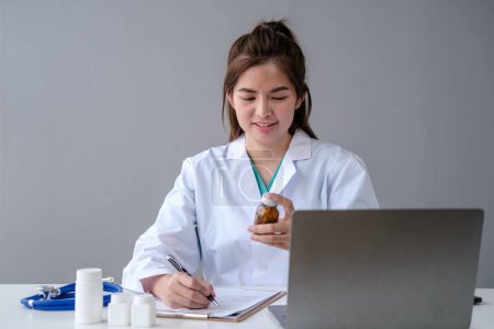 Téléchargez les photos : Souriant heureux médecin féminin travaillant avec ses pilules de documents pour ordinateur portable dans le bureau de l'hôpital de la chambre, Jeune médecin asiatique en uniforme médical blanc avec ordinateur portable de travail - en image libre de droit