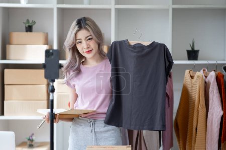 Photo for Young woman selling goods product clothes dress shirt online by smartphone live streaming video interaction internet online shop and talking to camera - Royalty Free Image