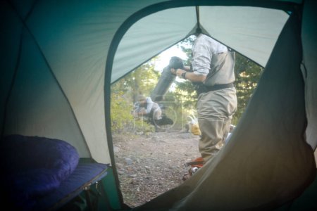 Photo for View from inside through an unzipped tent flap of two men preparing for the days activities outdoors on the campsite - Royalty Free Image
