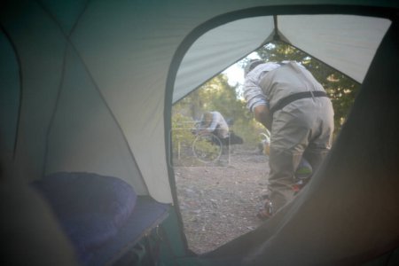 Photo for View through the unzipped flap from inside a tent of the campsite outside with two men preparing for the days activities - Royalty Free Image
