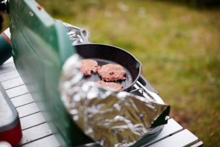 Cooking burger patties on a gas burner outdoors with foil wind guards set up on a small portable table while camping