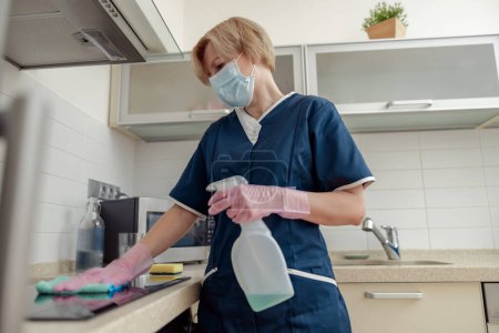 Foto de La señora de limpieza en máscara limpia la mesa de trabajo, usa trapo y productos de detergente efectivos en la cocina. - Imagen libre de derechos