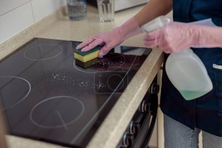 Foto de La señora de la limpieza en guantes protectores lava la cocina con esponja y aerosol. Concepto de servicio doméstico - Imagen libre de derechos