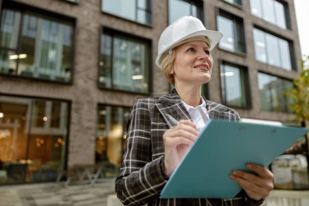 Foto de Mujer arquitecta en casco blanco sosteniendo tableta y mirando al lado en el fondo del edificio de la ciudad - Imagen libre de derechos