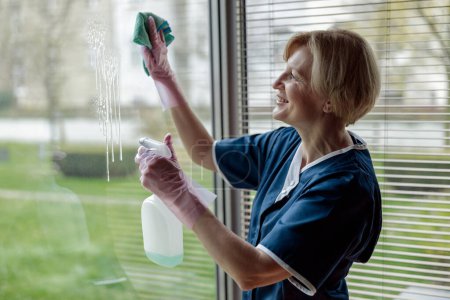 Téléchargez les photos : Femme de chambre en gants et fenêtre de nettoyage uniforme avec chiffon et spray nettoyant. Photo de haute qualité - en image libre de droit