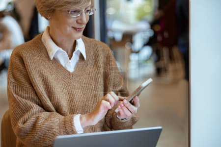 Photo for Middle aged woman in casual clothes works remotely using laptop and phone while sitting in coworking - Royalty Free Image