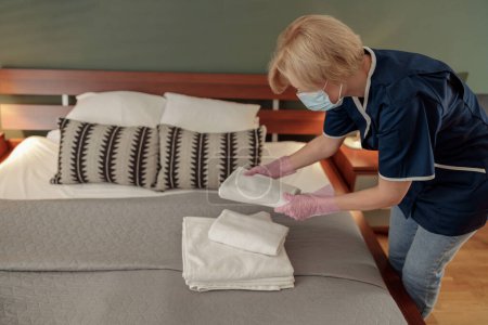 Téléchargez les photos : Femme de ménage dans le masque mettant pile de serviettes de bain blanches fraîches sur le drap de lit. Photo de haute qualité - en image libre de droit