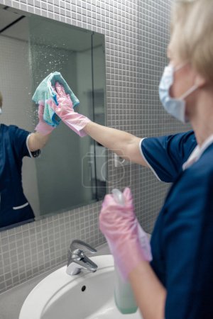 Téléchargez les photos : Nettoyage dame nettoie miroir dans la salle de bain pulvérisation détergent sur la surface. Concept de ménage - en image libre de droit