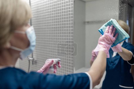 Téléchargez les photos : Nettoyage dame nettoie miroir dans la salle de bain pulvérisation détergent sur la surface. Concept de ménage - en image libre de droit