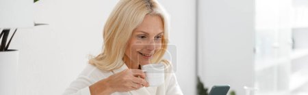 Photo for Caucasian mature lady holding a cup and looking at smartphone in modern spa center, beauty clinic - Royalty Free Image
