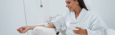 Téléchargez les photos : Femme en peignoir blanc boire de l'eau pendant la procédure médicale dans une clinique de beauté. Photo de haute qualité - en image libre de droit