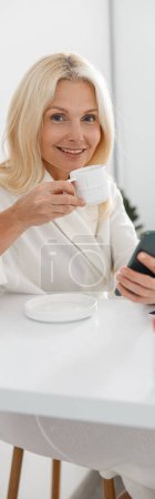 Foto de Senior hermosa mujer cliente beber café y el uso de teléfono inteligente en clínica de belleza, tiro vertical - Imagen libre de derechos
