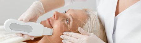 Téléchargez les photos : Cosmétologue professionnel fait nettoyage du visage par ultrasons pour belle femme blonde mature caucasienne heureuse dans une clinique moderne - en image libre de droit