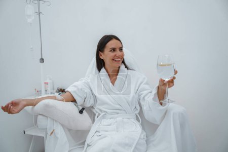 Foto de Mujer en albornoz blanco beber agua durante el procedimiento médico en la clínica de belleza. Foto de alta calidad - Imagen libre de derechos