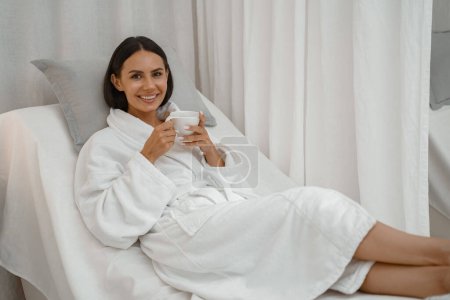 Foto de Mujer bonita feliz en albornoz blanco disfrutando mientras descansa con una taza de café en el salón de spa - Imagen libre de derechos