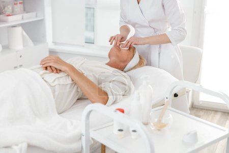 Foto de Hermosa médico profesional mujer está limpiando una piel de mujer rubia caucásica madura en el centro de cosmetología luz moderna - Imagen libre de derechos