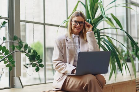 Smiling adult business woman working laptop while sitting in modern office. Blurred background