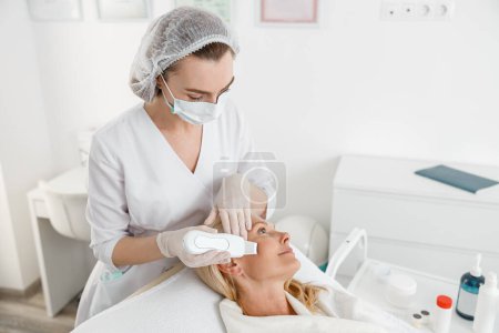 Téléchargez les photos : Cosmétologue féminine travaillant avec une patiente utilisant un nettoyeur de visage ultrasonique effectuant une procédure de beauté dans une clinique moderne - en image libre de droit