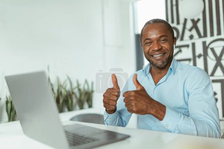 Téléchargez les photos : Homme d'affaires africain souriant montrant les doigts levés alors qu'il était assis au bureau. Photo de haute qualité - en image libre de droit